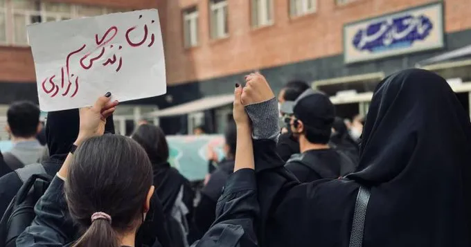 An undated photo captures a protest at the Art University of Tehran. Two women, one without a hijab and the other wearing one, stand united hand-in-hand. Their shared message is powerfully displayed on a sign that reads, "Women: Life, Freedom."